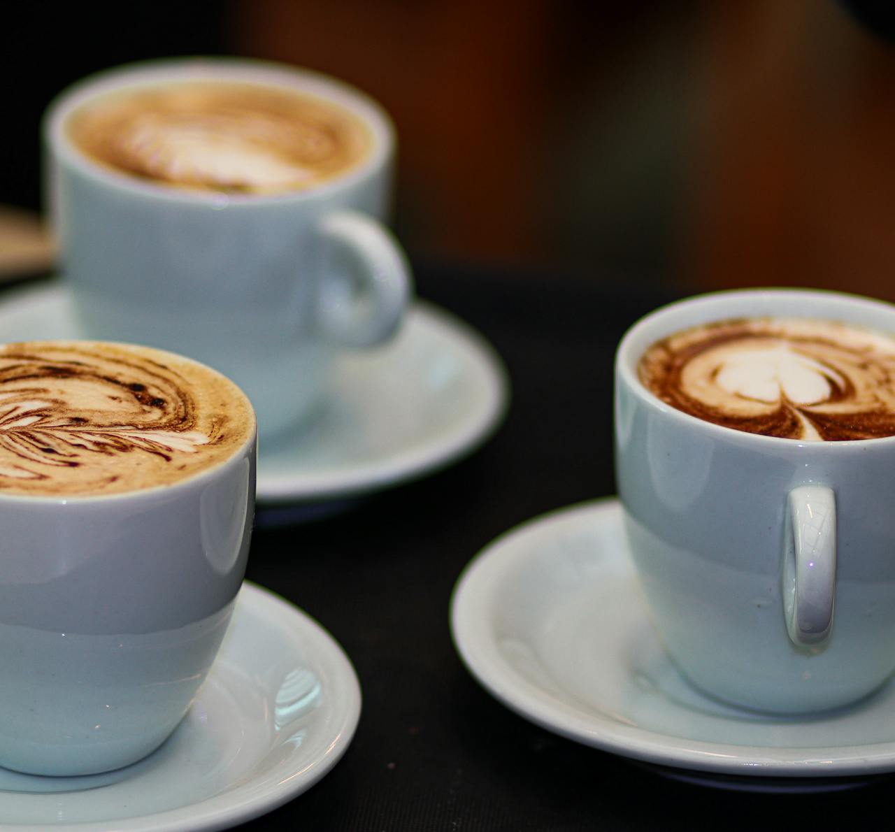 Three cappuccinos with intricate latte art in white cups on saucers, showcasing coffee artistry.